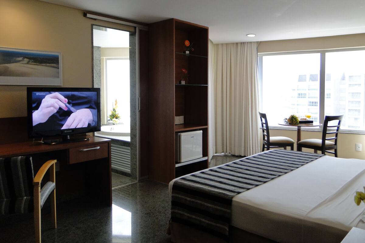 A hotel room with a bed, a desk with a television, a bookshelf, a window with curtains, and a view of buildings.