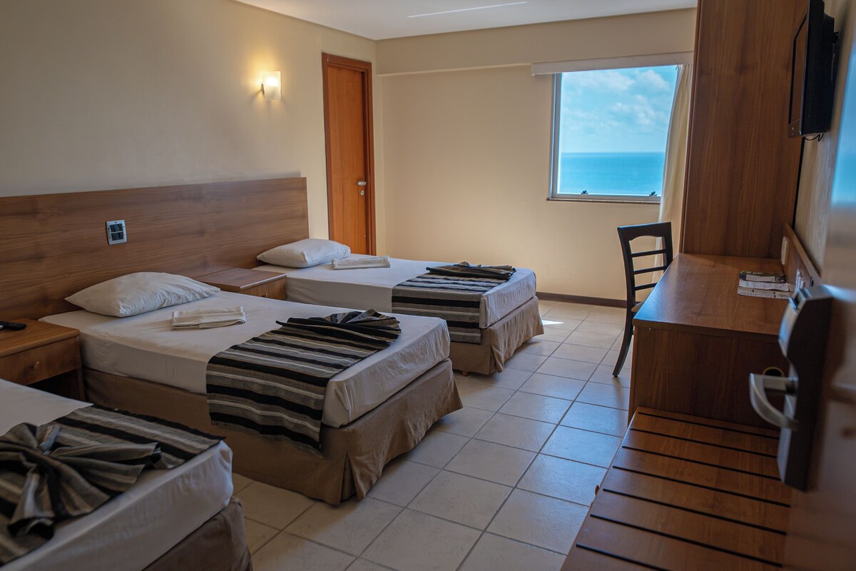 A hotel room with two beds, a desk, a chair, a TV, and a window with a view of the sea.