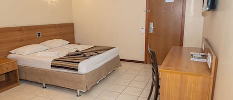 A hotel room with a bed, a desk, and a chair. There is a wall-mounted air conditioner and a light fixture above the bed.