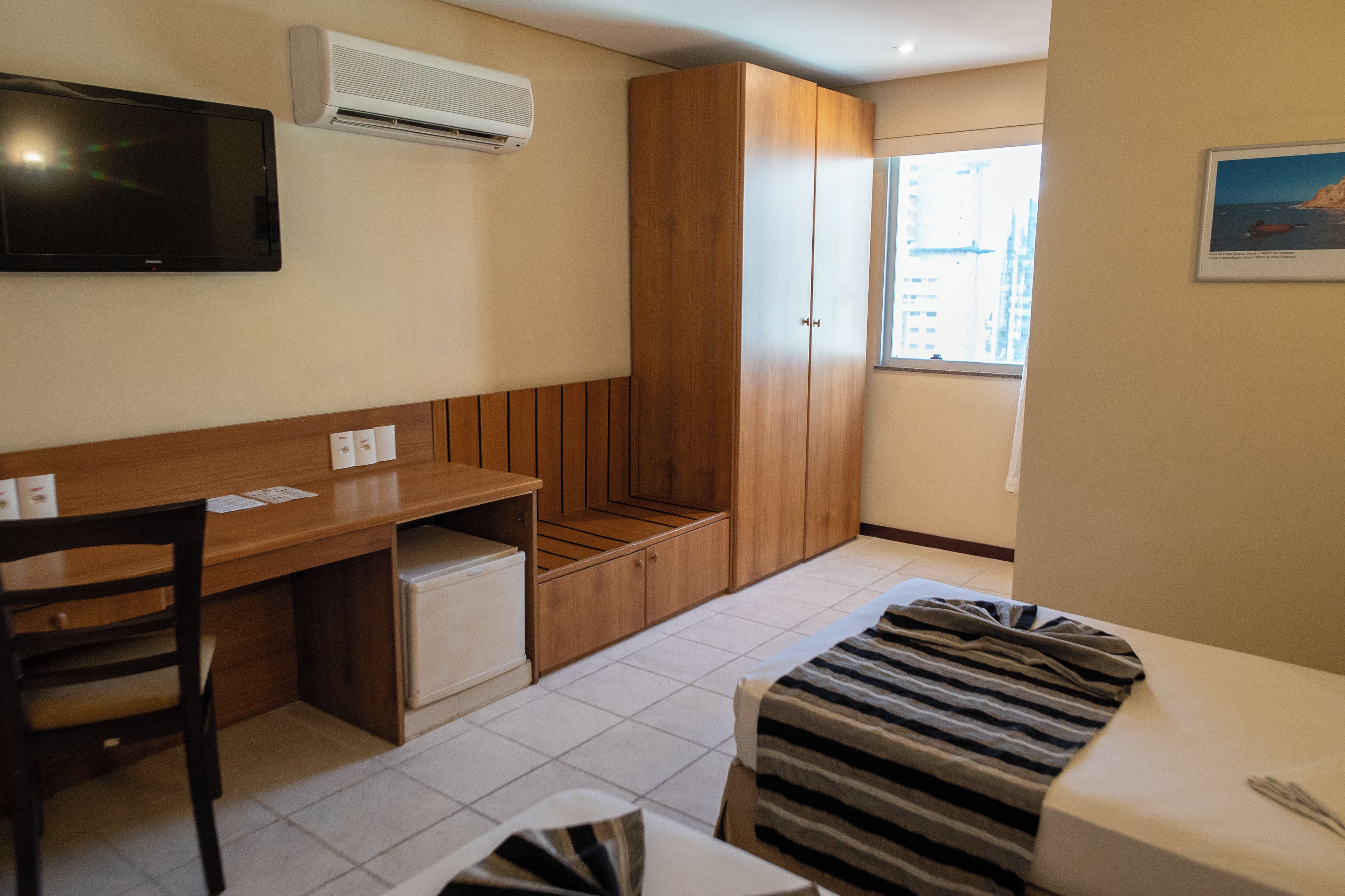 Une chambre d’hôtel compacte comprenant un coin cuisine, un bureau en bois, un petit réfrigérateur, un lit avec une couverture à rayures, un téléviseur fixé au mur et une fenêtre donnant sur le paysage urbain.
