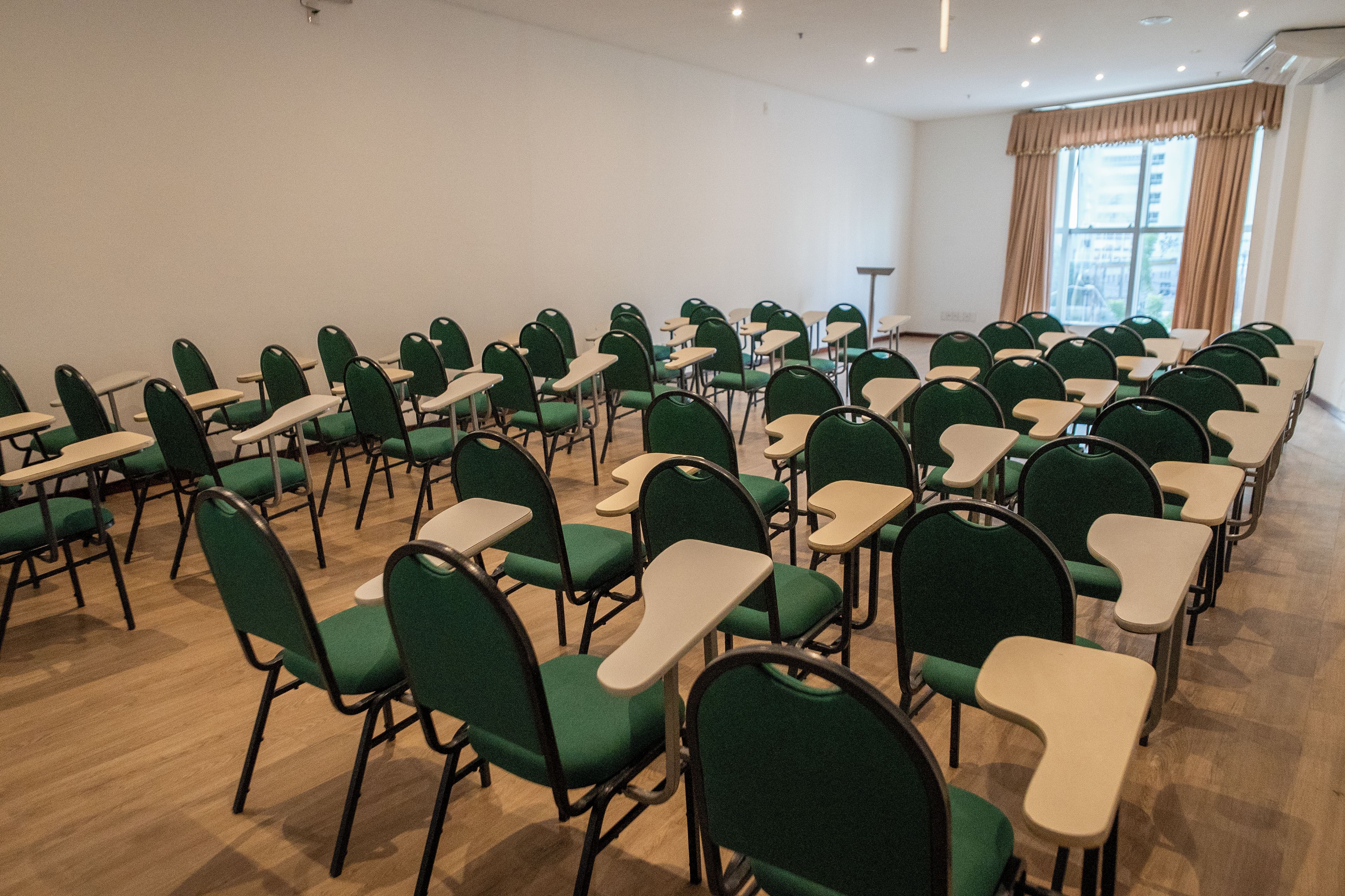 Une salle de conférence avec des rangées de chaises et de tables vertes, une grande fenêtre avec des rideaux et un plafond doté d’un éclairage encastré.