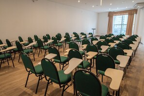 Een congrescentrum met rijen groene stoelen en tafels, een groot raam met gordijnen en een plafond met inbouwverlichting.
