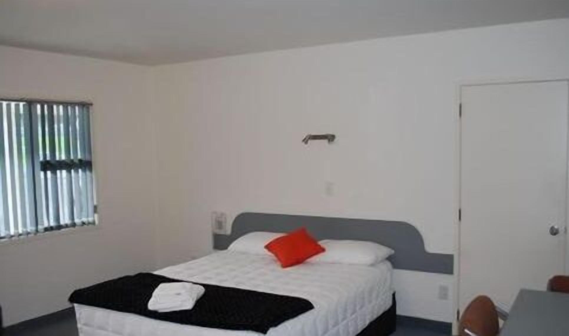 Una cama individual con una colcha blanca y negra, un almohadón rojo y una toalla blanca. Se ve una ventana con persianas a la izquierda y una puerta a la derecha.