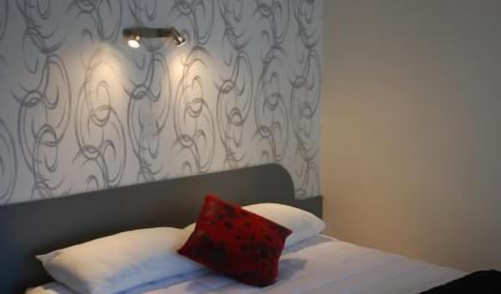 A bed with a white headboard and a red pillow, a wall with a patterned design, and a light fixture above the bed.