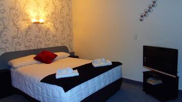 A neatly made bed with white linens and a red pillow, a black blanket, and two folded towels. A flat-screen TV is on a stand beside the bed.
