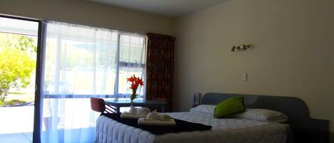 A bedroom with a bed, a small table with a vase of flowers, a chair, and a view of the outdoors through a large window.