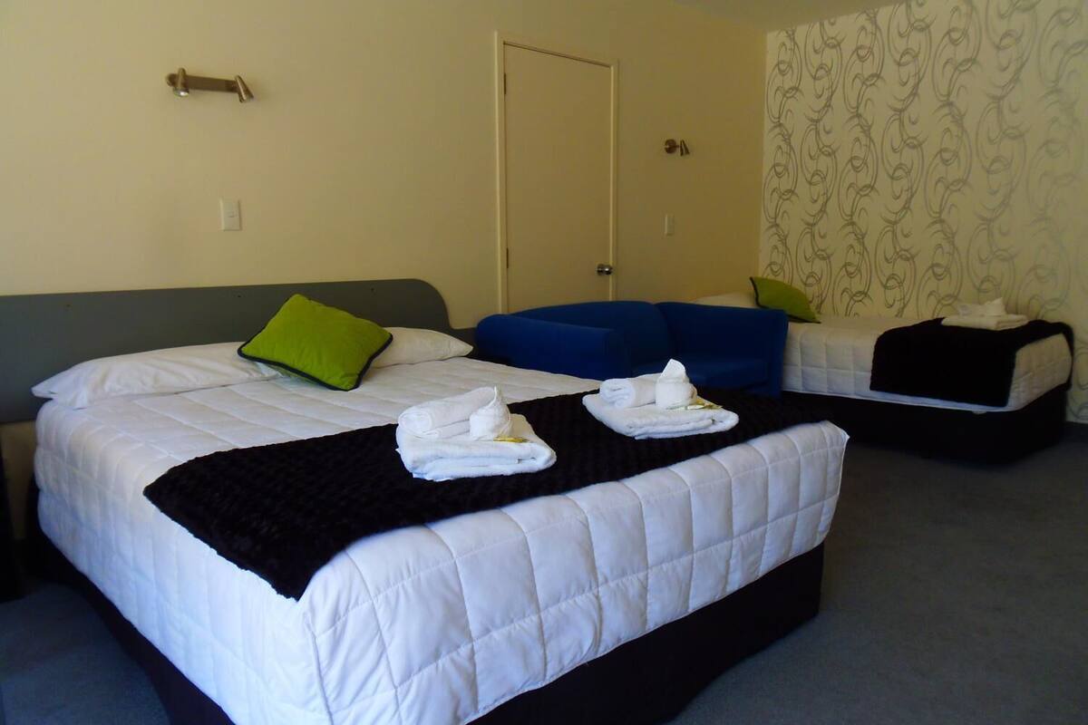 Ein Hotelzimmer mit zwei Betten, jeweils mit schwarzer, gesteppter Tagesdecke und weißen Bettwäsche. Auf den Betten liegen grüne und weiße Kissen. Das Zimmer hat eine Tür und die Wände sind mit einem gemusterten Wandpapier versehen.