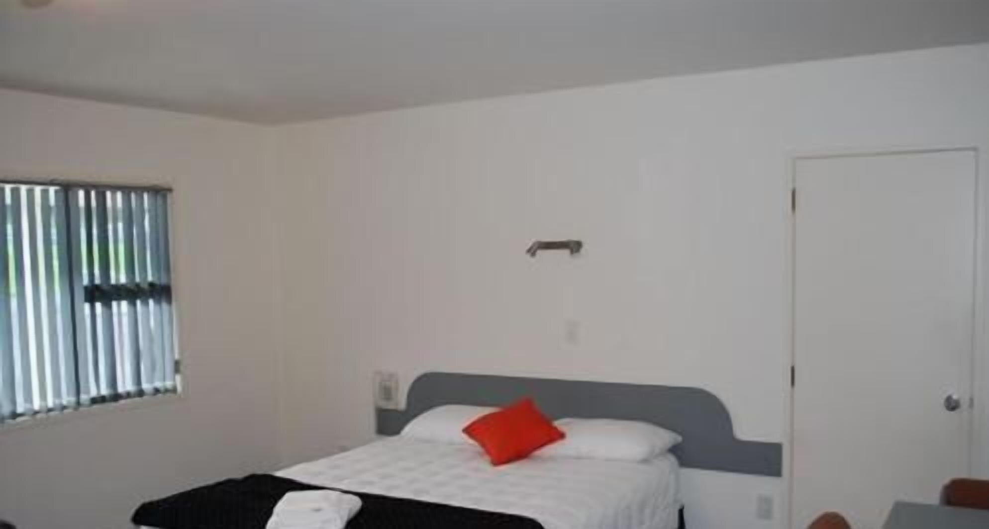A hotel room with a bed, a window with blinds, a door, and a small bedside table.