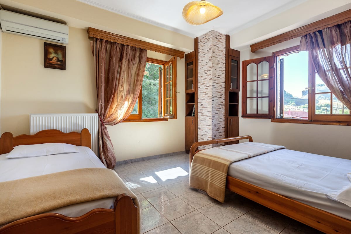 A room with two wooden beds, a brick pillar, a window with curtains, and a ceiling fan.