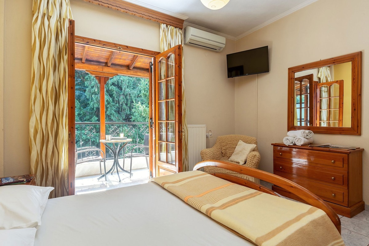 A bedroom with a bed, a wooden dresser, a chair, a television, and a balcony with a table and chairs.