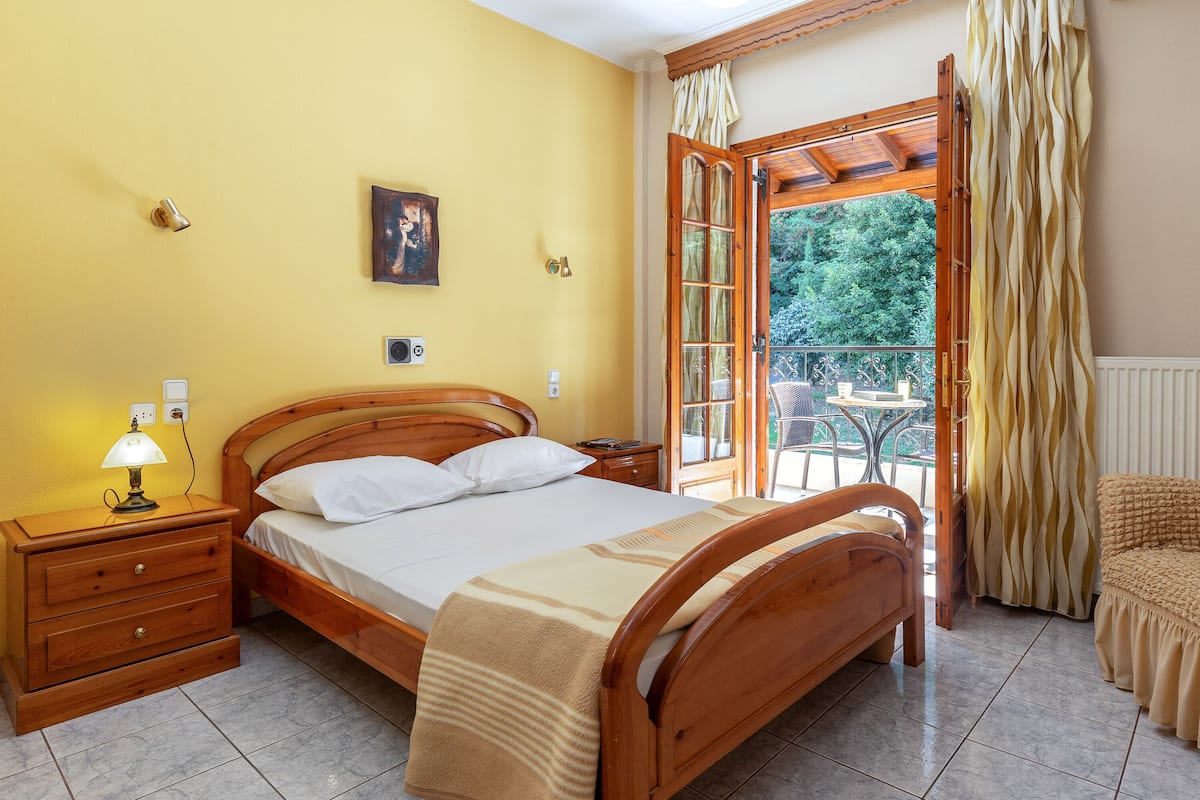 A bedroom with a wooden bed, a nightstand with a lamp, a window with curtains, and a view of greenery outside.