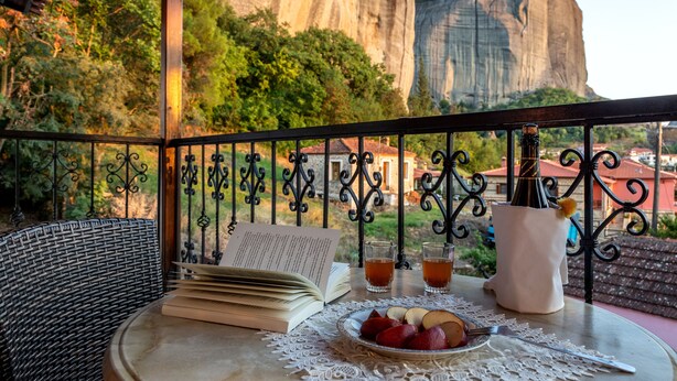 Un balcón con una mesa dispuesta para dos personas, que incluye un libro, dos vasos de una bebida, un plato de fruta y un jarrón con una flor.