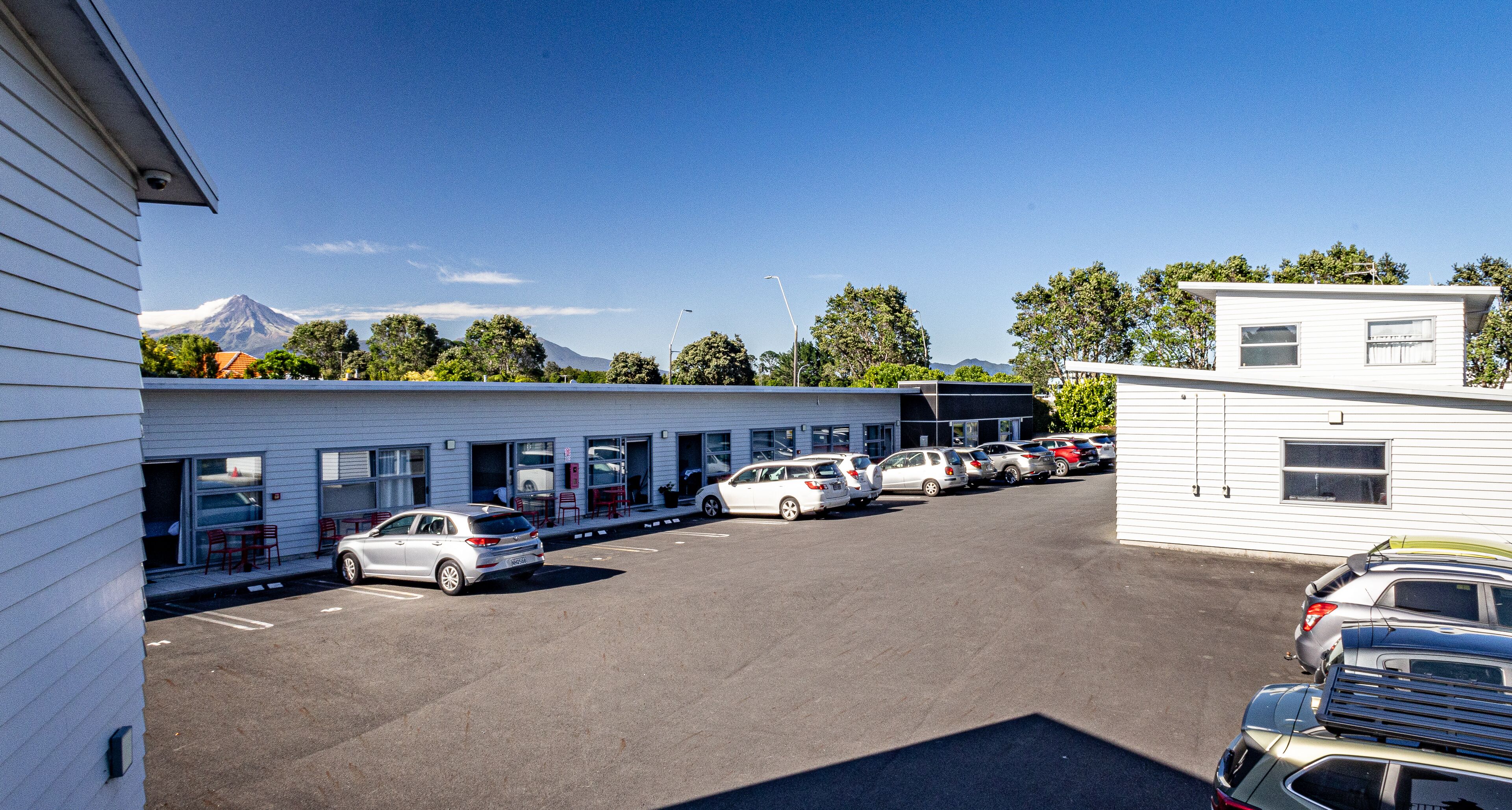 Un parking avec plusieurs voitures, un bâtiment à l’extérieur blanc et une montagne en arrière-plan.