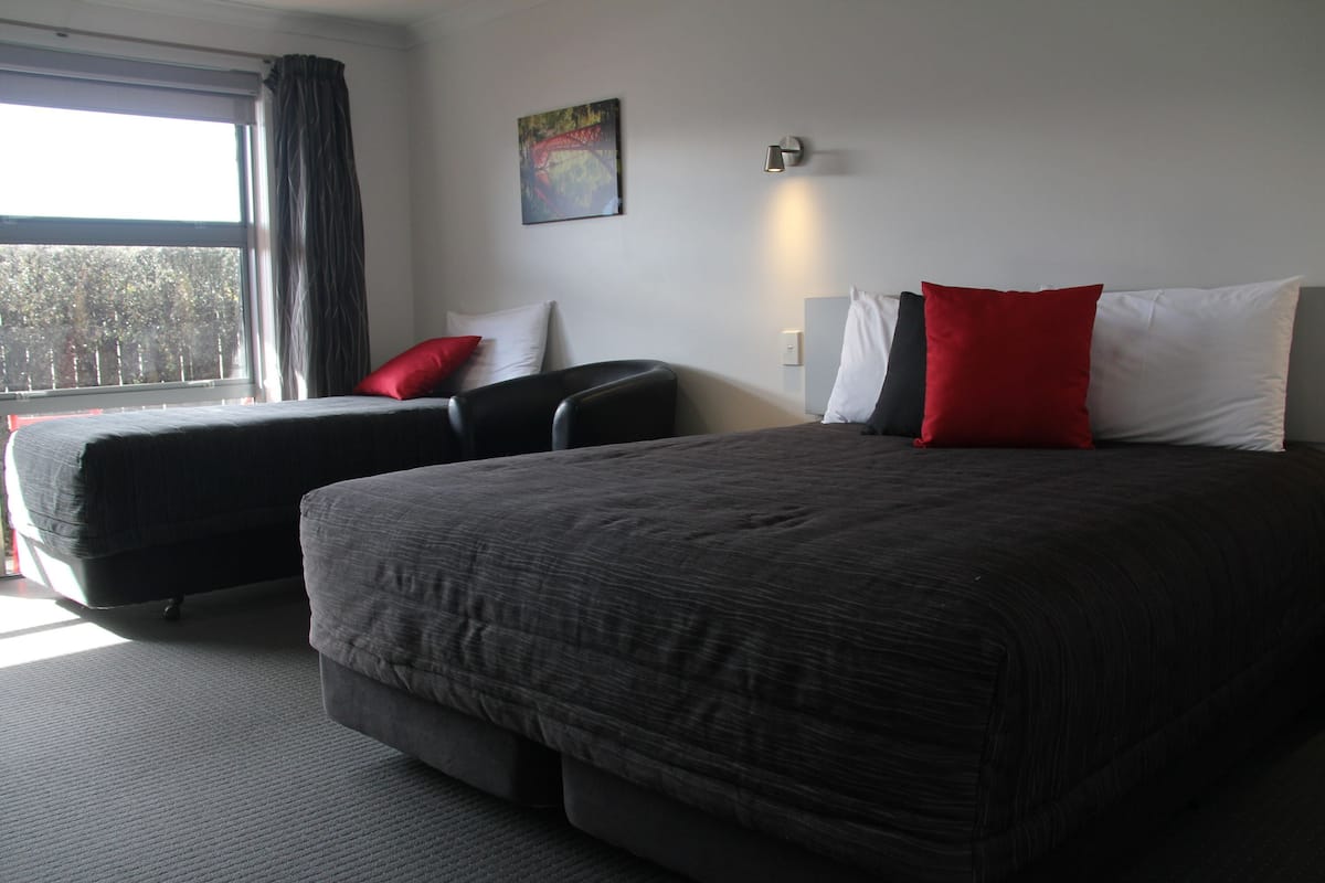 Una habitación de hotel con una cama grande, una cama más pequeña, una silla, una ventana con cortinas, un cuadro en la pared y una lámpara de pared.