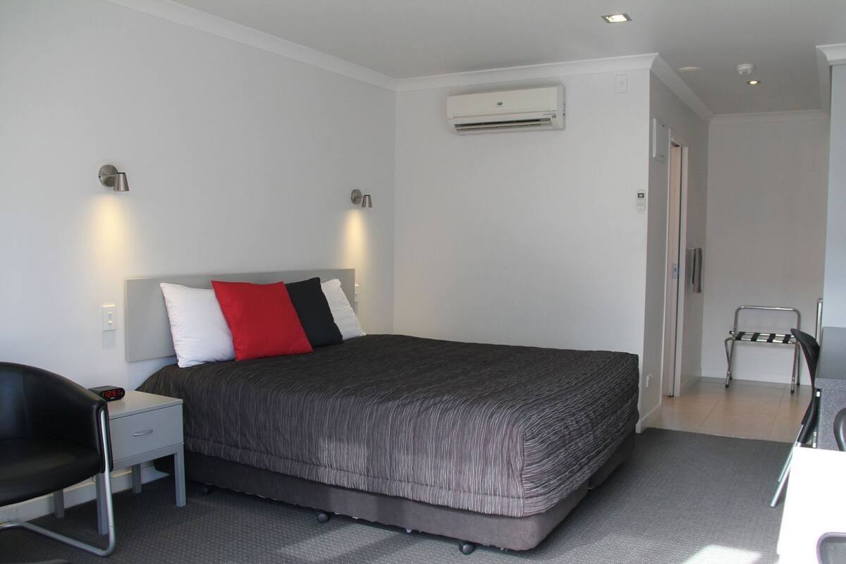 Una habitación de hotel moderna con una cama, un escritorio con una silla, un equipo de aire acondicionado y una silla en la esquina.