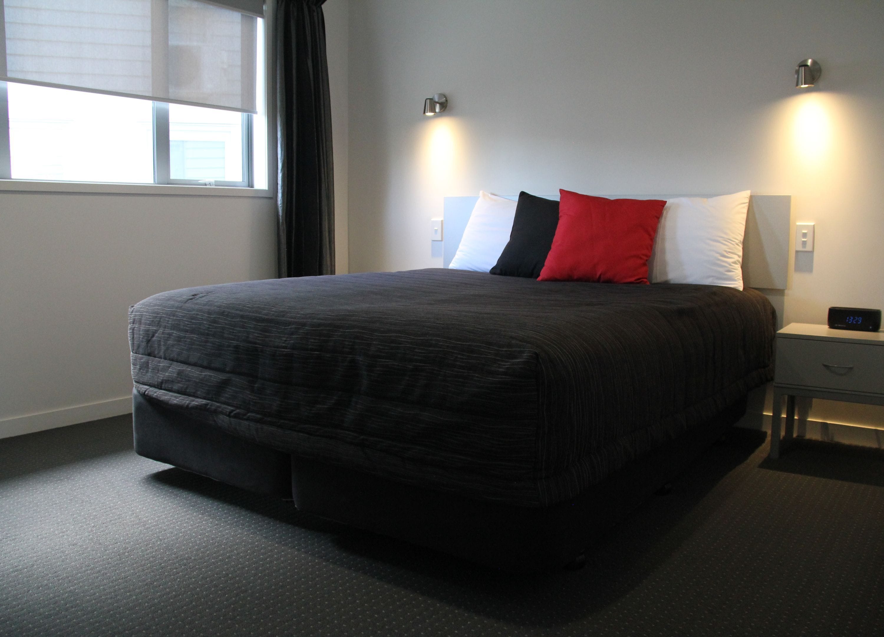 A neatly made bed with a dark quilt, a bedside table with a digital clock, and a window with blinds.