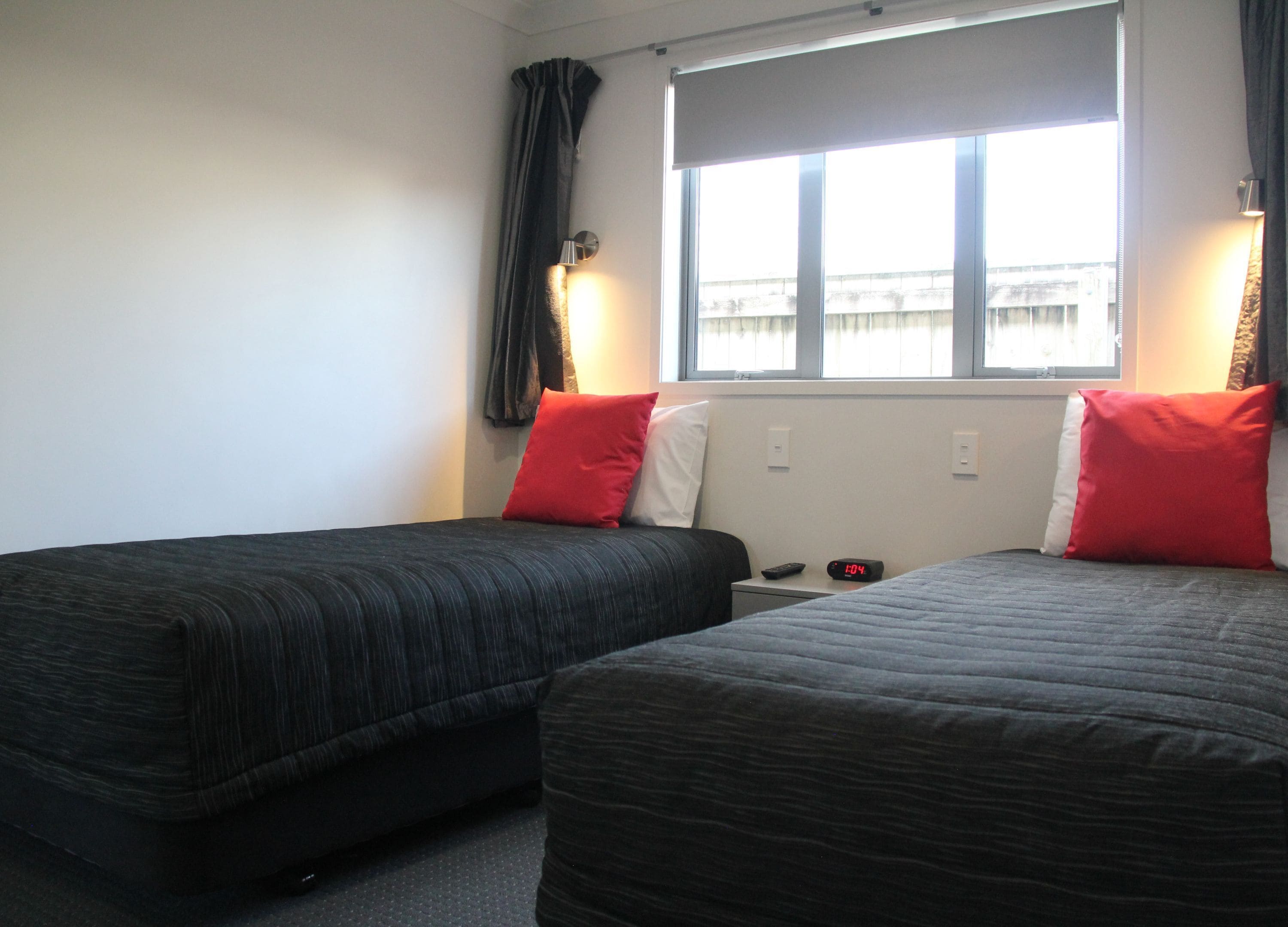Ein Hotelzimmer mit zwei Betten, jeweils mit schwarzer Bettdecke und roten Kissen. Ein Fenster bietet Blick auf Gebäude, und ein Nachttisch ist mit einer Uhr und einer Fernbedienung ausgestattet.