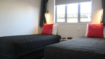 Ein Hotelzimmer mit zwei Betten, jeweils mit schwarzer Bettdecke und roten Kissen. Ein Fenster bietet Blick auf Gebäude, und ein Nachttisch ist mit einer Uhr und einer Fernbedienung ausgestattet.