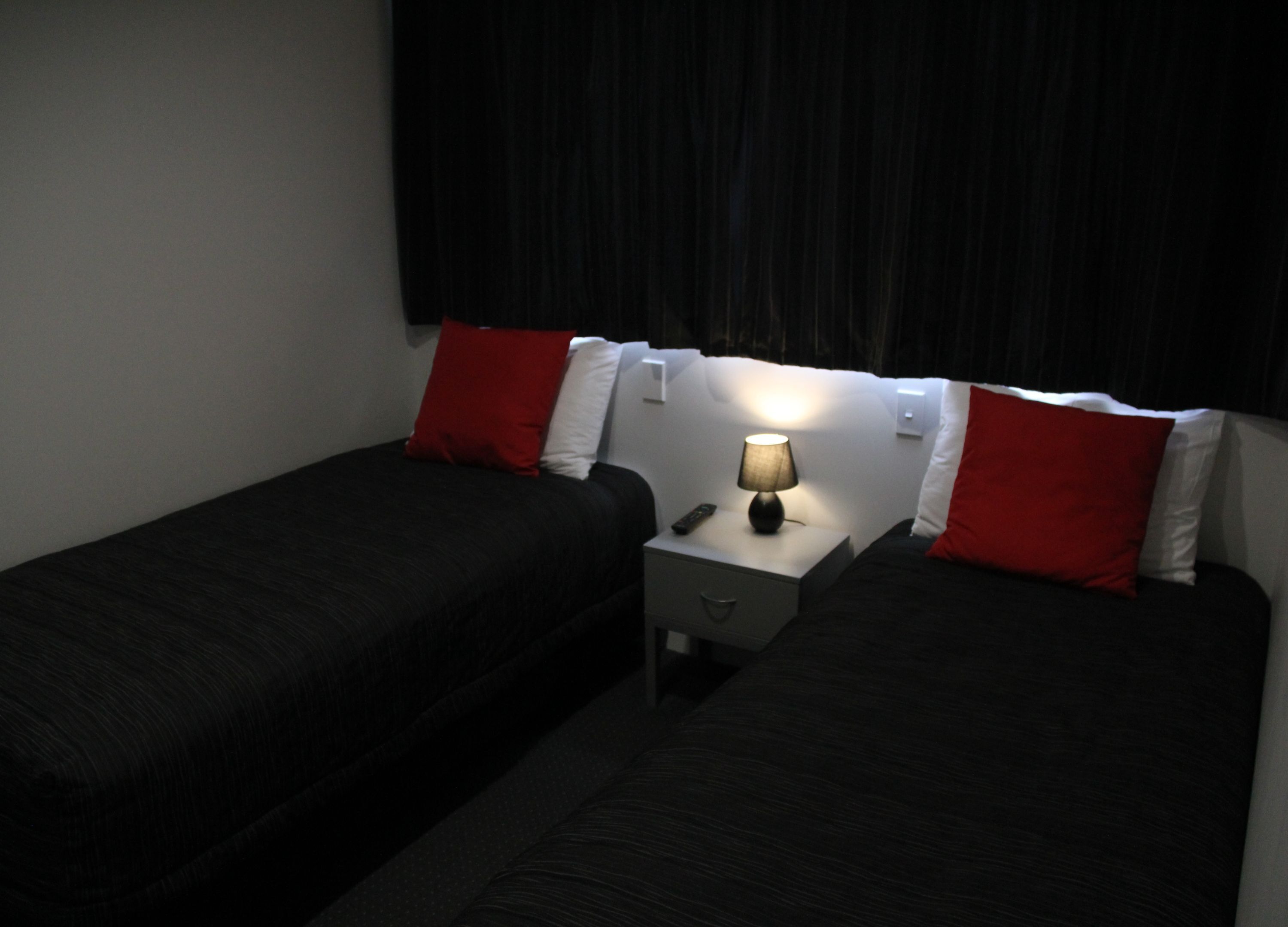 Una habitación con dos camas individuales, cada una con cubrecama negro y almohadas rojas. Entre las camas, un mesita de noche con una lámpara.