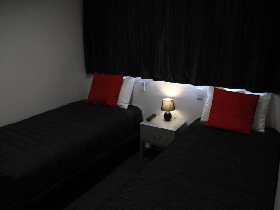 Une chambre avec deux lits simples, chacun recouvert d’une couverture noire et agrémenté d’oreillers rouges. Une table de chevet avec une lampe se trouve entre les deux lits.