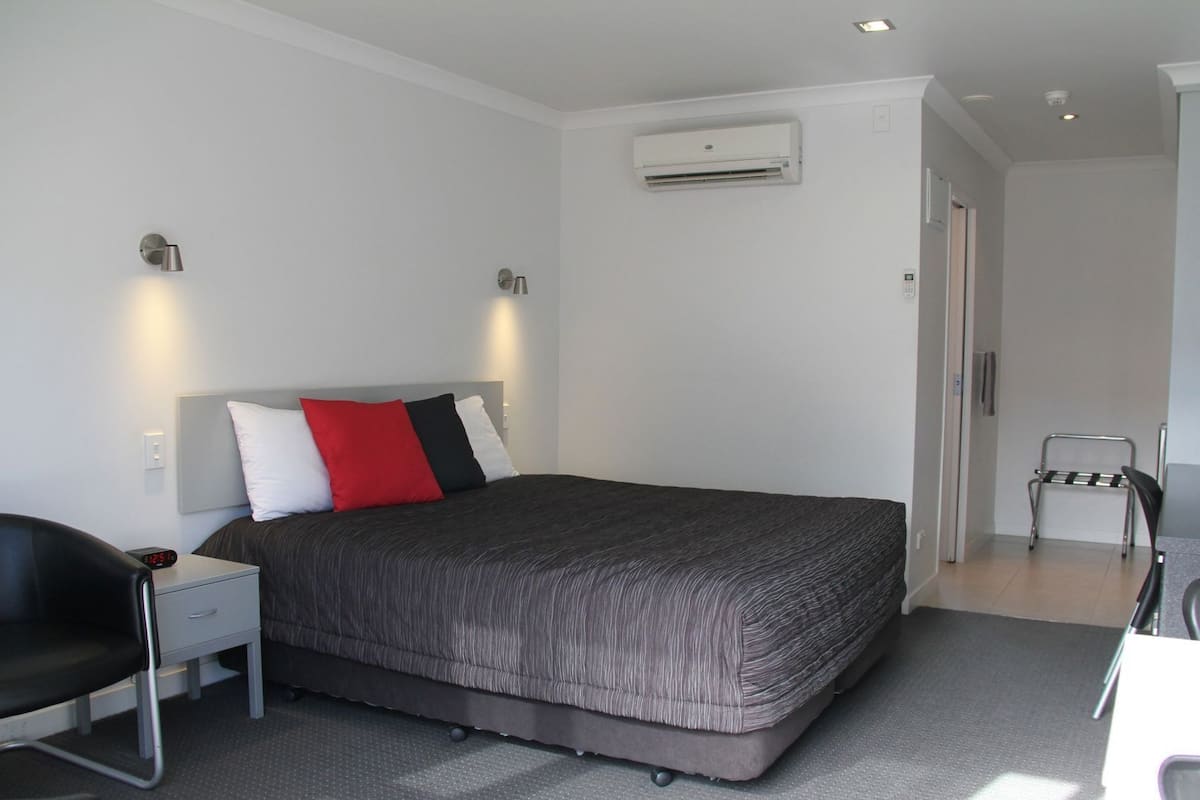Una habitación de hotel moderna con una cama, un escritorio con una silla, un equipo de aire acondicionado y una silla en la esquina.