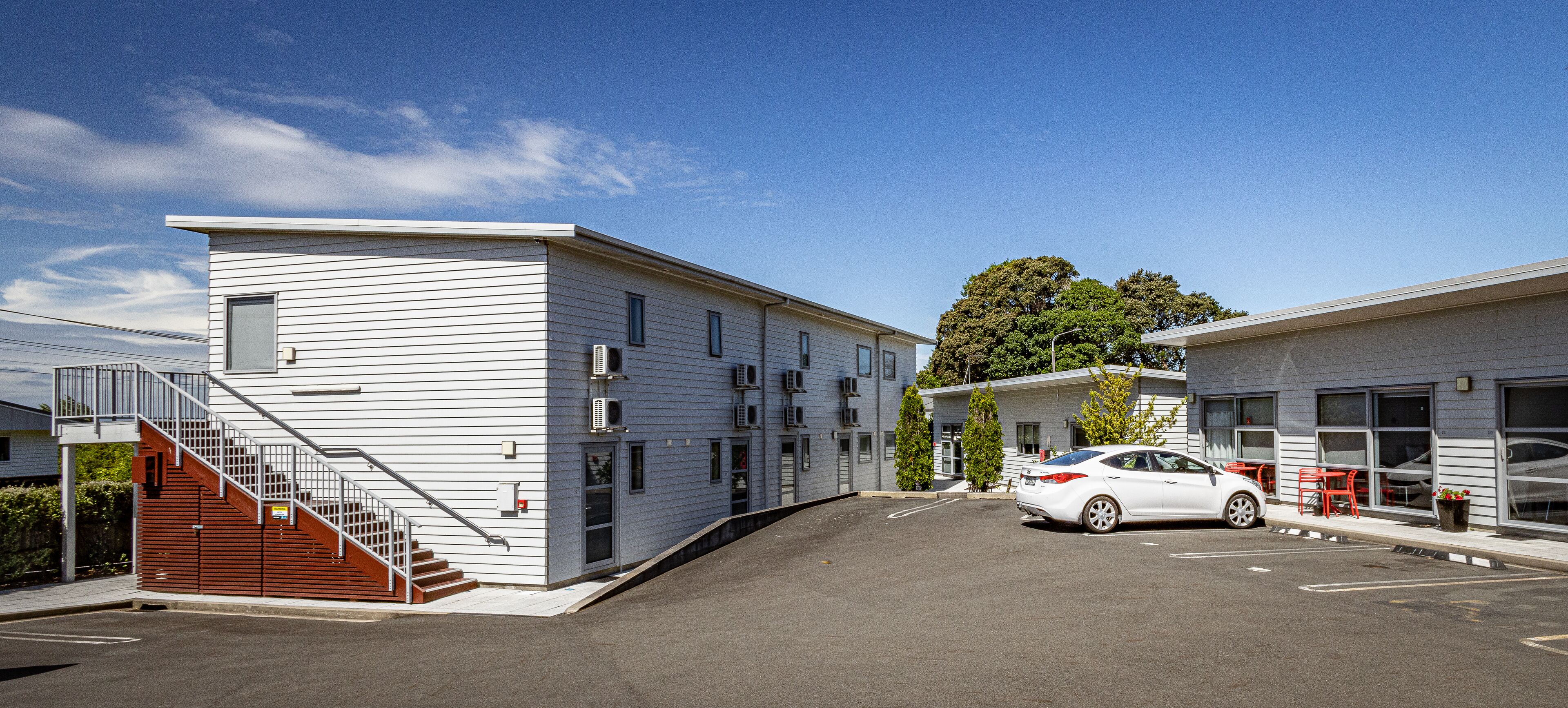 A modern building with a staircase, a white car parked in front, and a tree in the background.