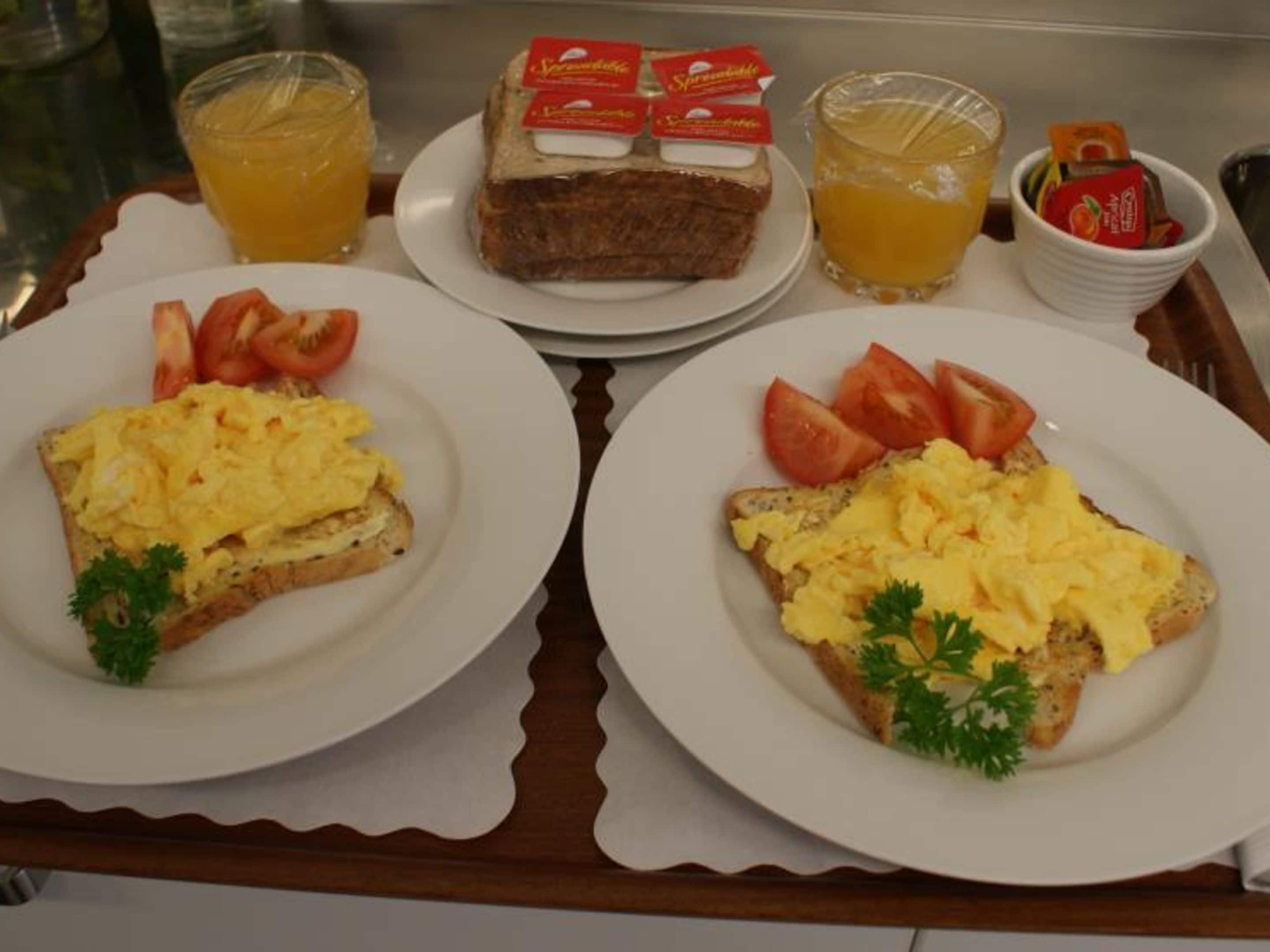 A breakfast tray with toast topped with scrambled eggs, tomatoes, and parsley, accompanied by two glasses of orange juice and a plate of bread with jam.