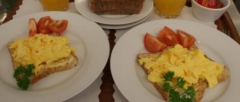 A breakfast tray with toast topped with scrambled eggs, tomatoes, and parsley, accompanied by two glasses of orange juice and a plate of bread with jam.