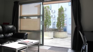 Un salon moderne avec un canapé en cuir noir, une table basse blanche et une porte-fenêtre coulissante donnant sur un balcon agrémenté de plantes en pot et sur lequel on aperçoit un immeuble en arrière-plan.