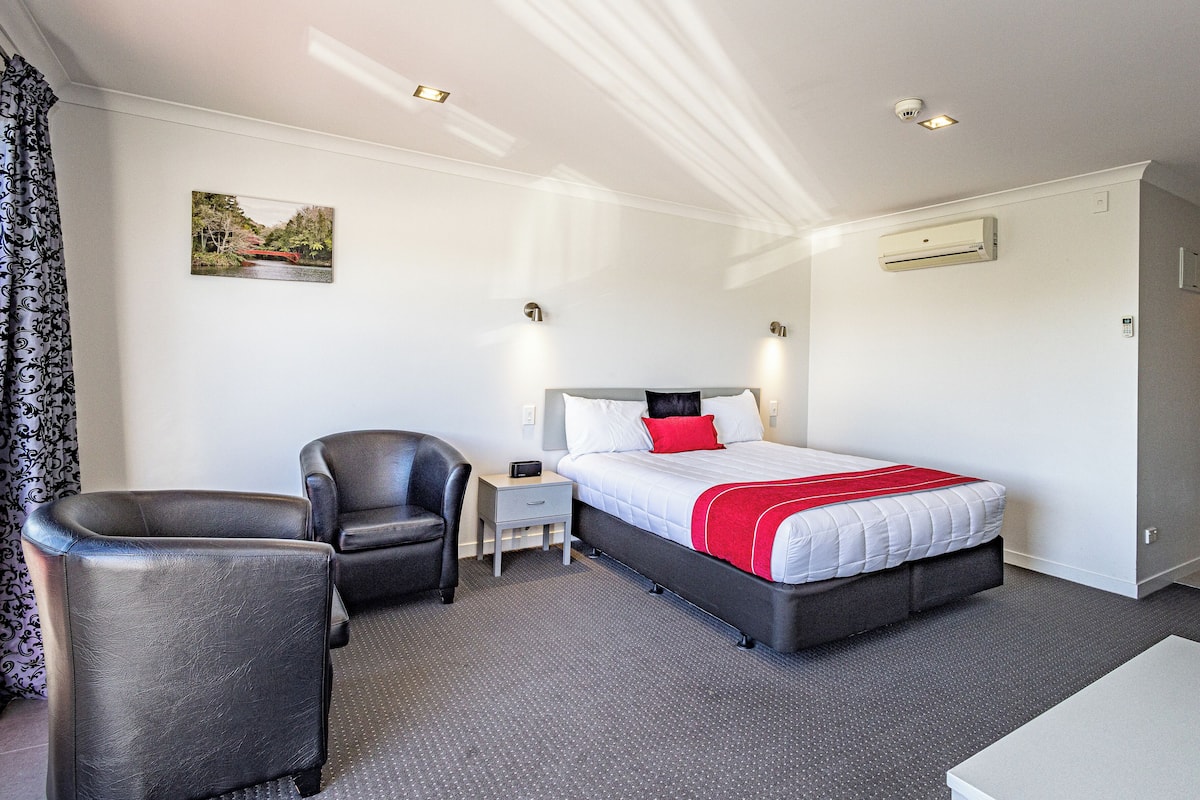 Una habitación de hotel con cama, sillón, mesita de noche, un cuadro en la pared y una puerta.