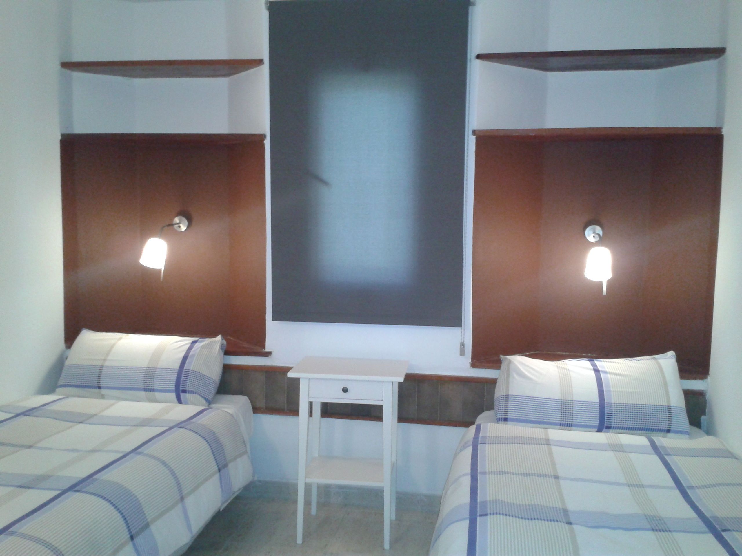 A hotel room with two beds, a small table, and wall-mounted lights.