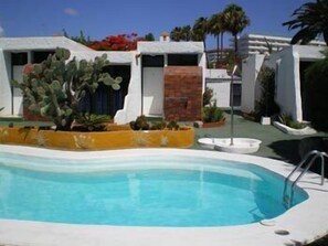 A swimming pool with a clear blue water, surrounded by a white pool deck. There are plants and a cactus in the foreground. - Bungalows Tenesoya - Caters to Gay Men (San Bartolomé de Tirajana)