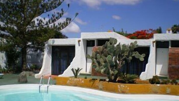 A pool area with a clear blue pool, surrounded by white buildings with blue doors, and a cactus plant in the foreground.