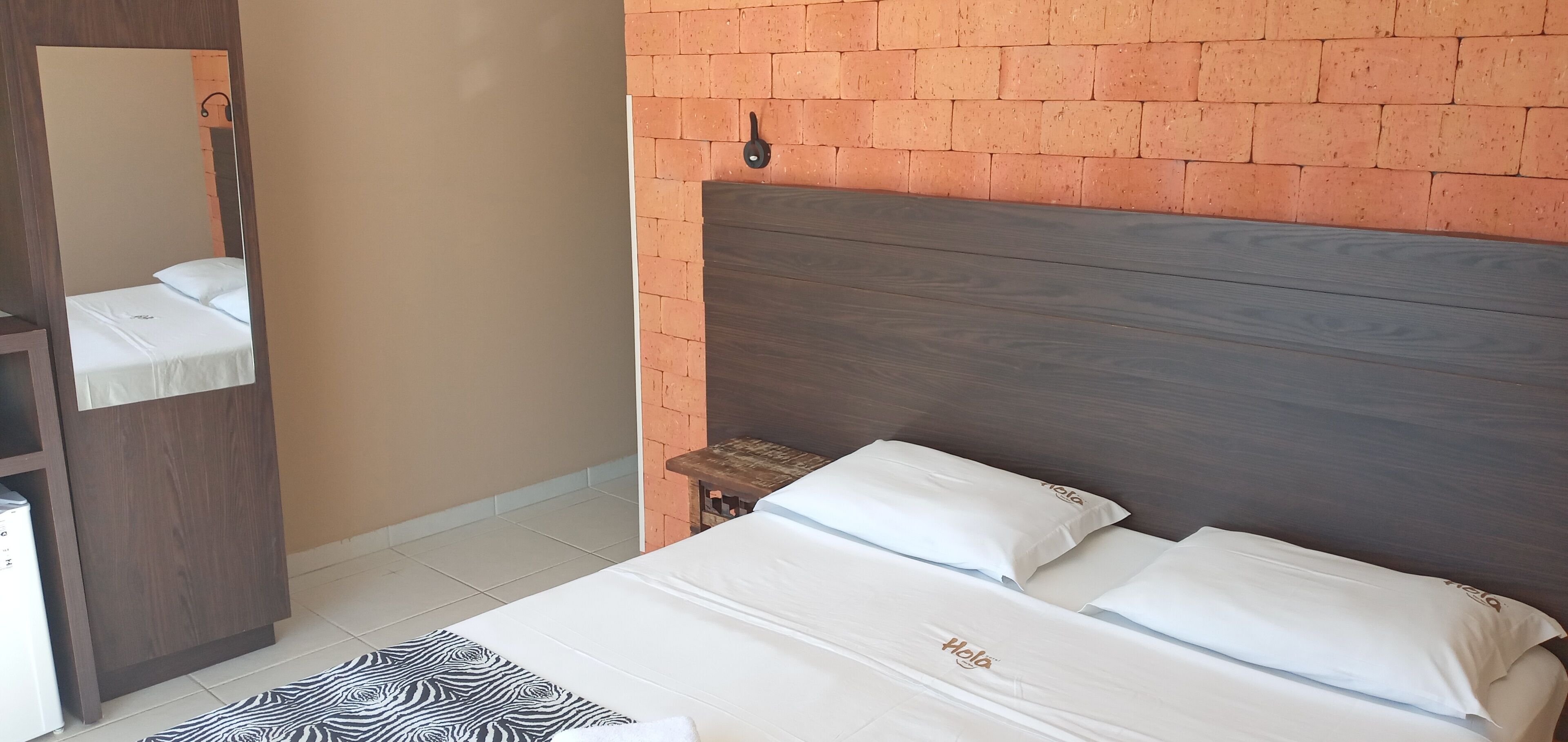 A hotel room with a brick-patterned wall, a wooden headboard, a bed with white bedding, a small wooden nightstand, and a mirror.