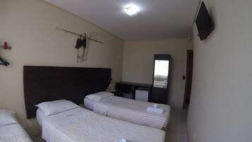 Quarto de hotel com três camas de solteiro, cabeceira escura, televisão de tela plana e pequena escrivaninha com espelho.