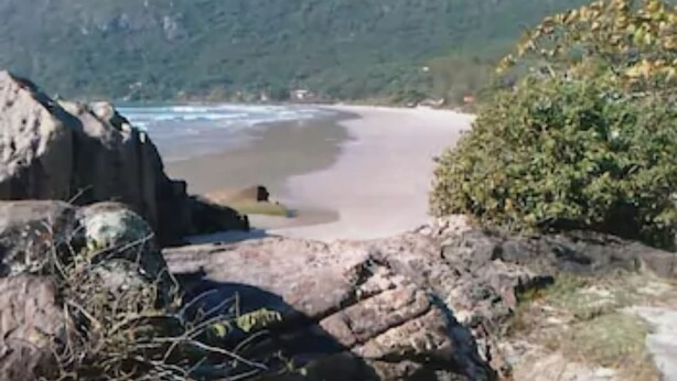 A coastal landscape with a beach, rocky outcrops, and a mountain in the background. - Hotel Hola (Florianópolis)