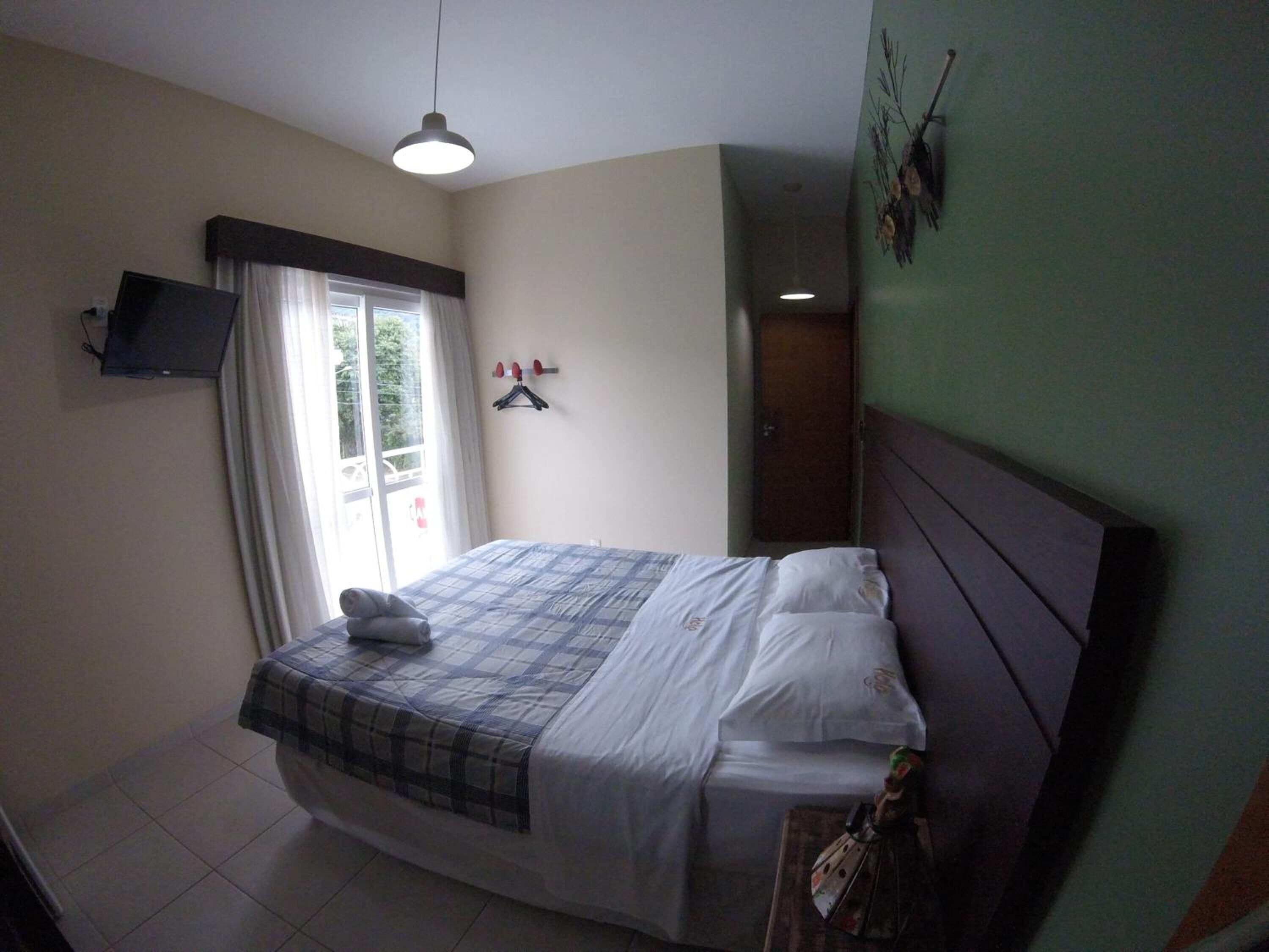 A bedroom with a bed, a flat-screen TV mounted on the wall, a window with curtains, and a door.