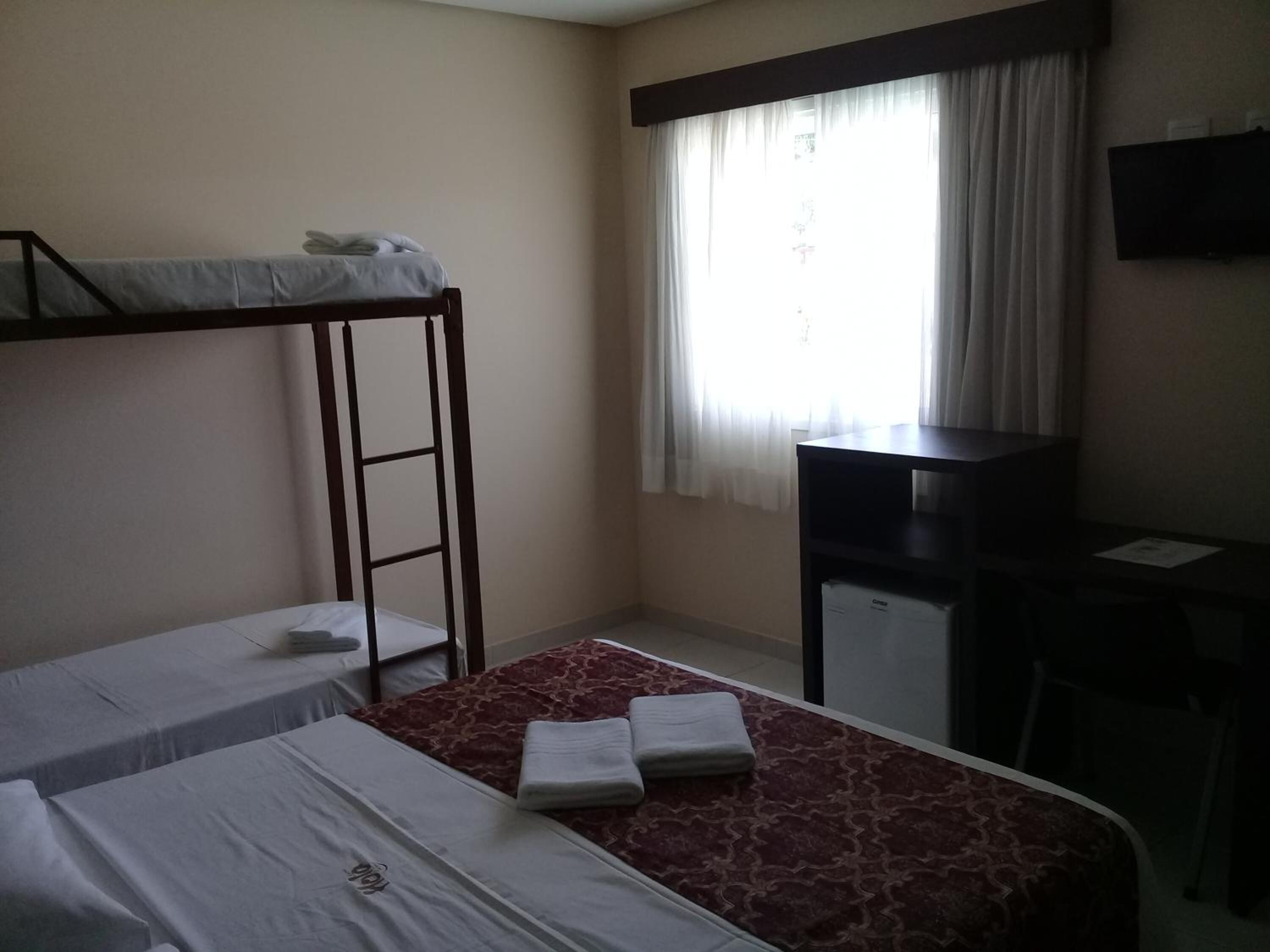 A room with two beds, one with a bunk, a desk, a chair, and a TV on the wall.