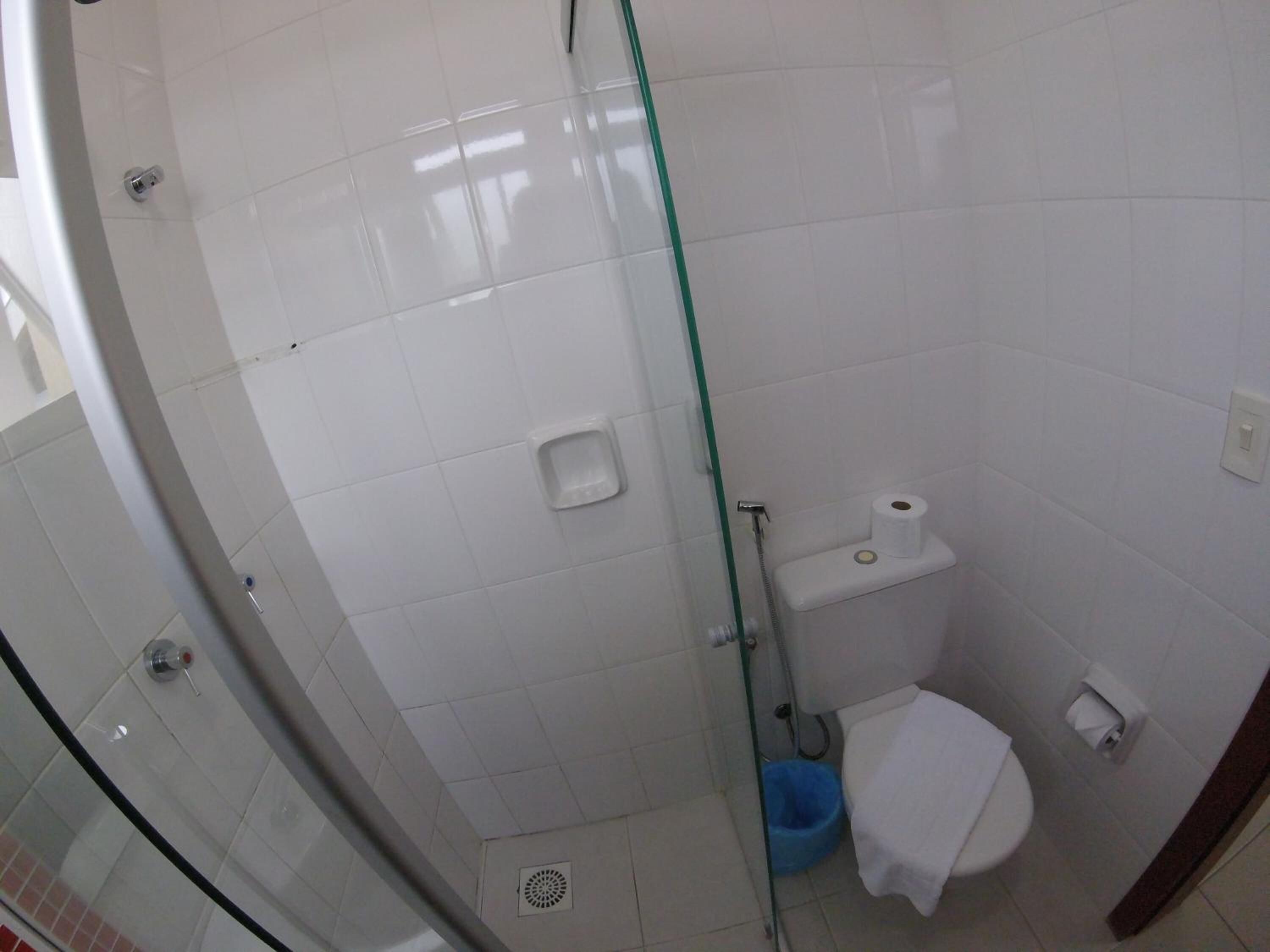 Un baño pequeño y funcional con azulejos blancos, mampara de vidrio, inodoro y un cubo de basura azul.