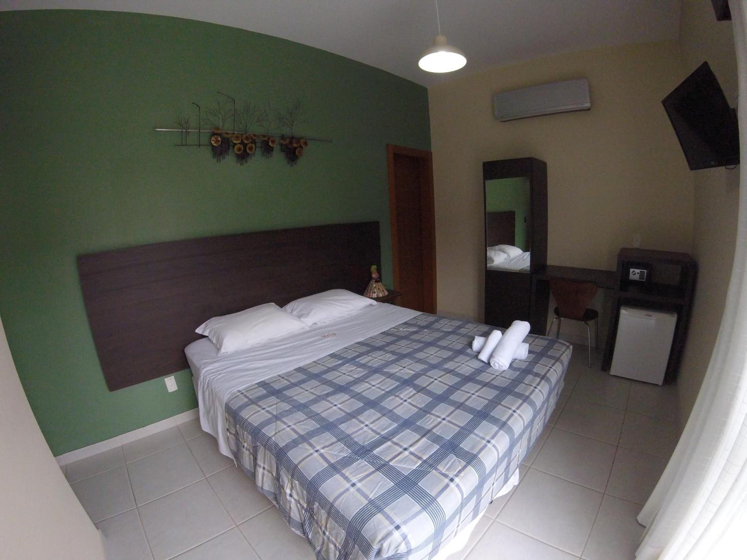 A hotel room with a bed, a small desk, a mirror, and a wall-mounted air conditioner.