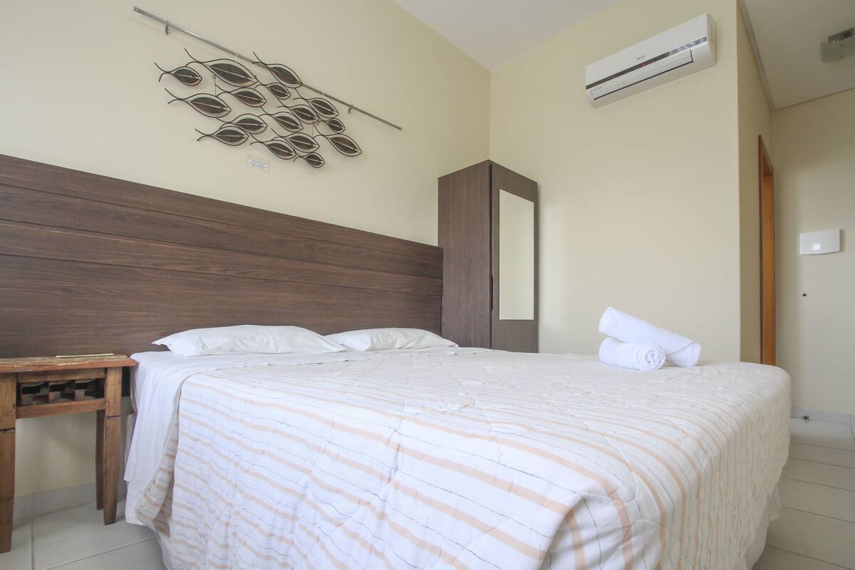 A neatly made bed with a striped bedspread, a wooden nightstand, a wall-mounted air conditioner, and a decorative wall art piece.