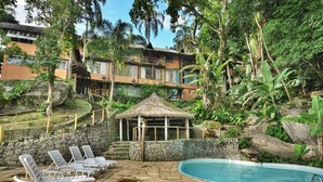 A pool area with lounge chairs, a thatched-roof structure, and a stone-paved deck. - Porto Pousada Saco da Capela (Ilhabela)