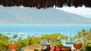 A table with a meal and drinks overlooking a coastal view with boats and hills. - Porto Pousada Saco da Capela (Ilhabela)
