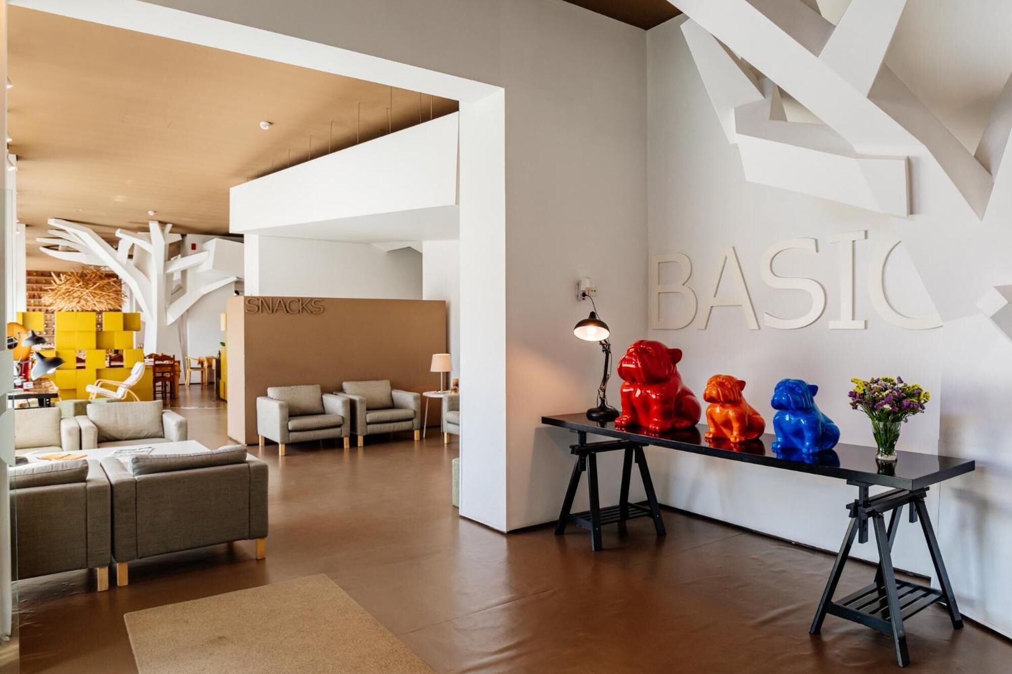 A modern hotel lobby with a central seating area, a 'SNACKS' counter, and a 'BASIC' sign on the wall.