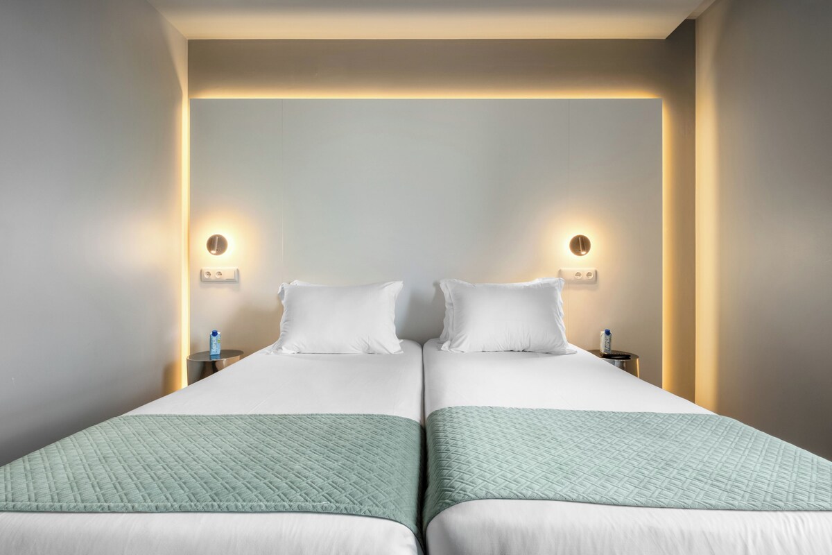 A double bed with a quilted green blanket, two bedside tables with a bottle of water, and wall-mounted light fixtures.