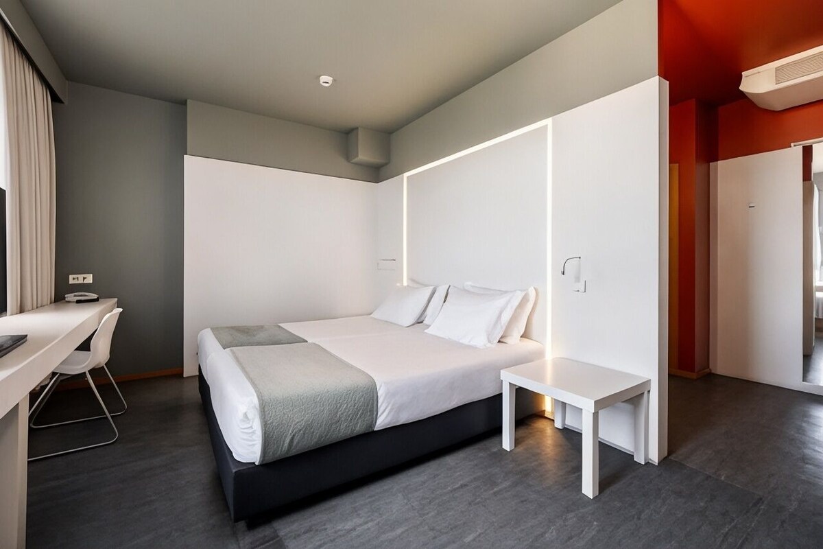 A modern hotel room with a bed, bedside table, desk, and chair.