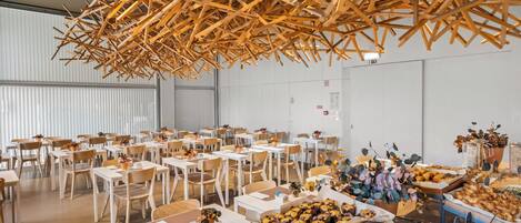 Een ruime eetruimte met meerdere tafels en stoelen, een houten plafondconstructie en diverse gerechten gepresenteerd op tafels.