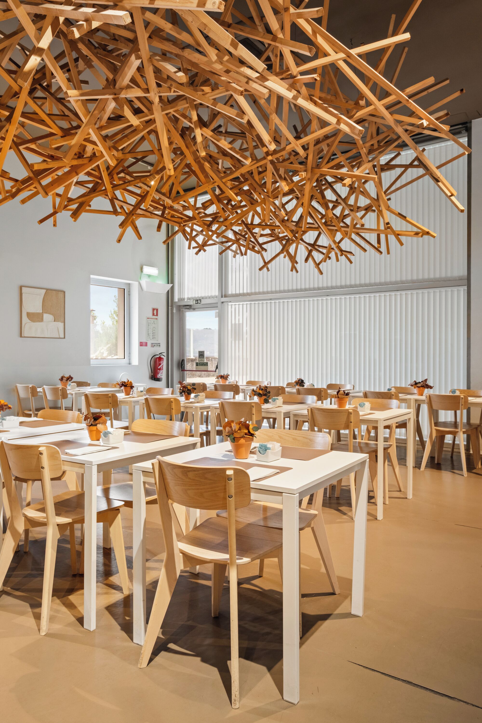 Un comedor con mesas y sillas de madera, una estructura colgante de madera y ventanas grandes.