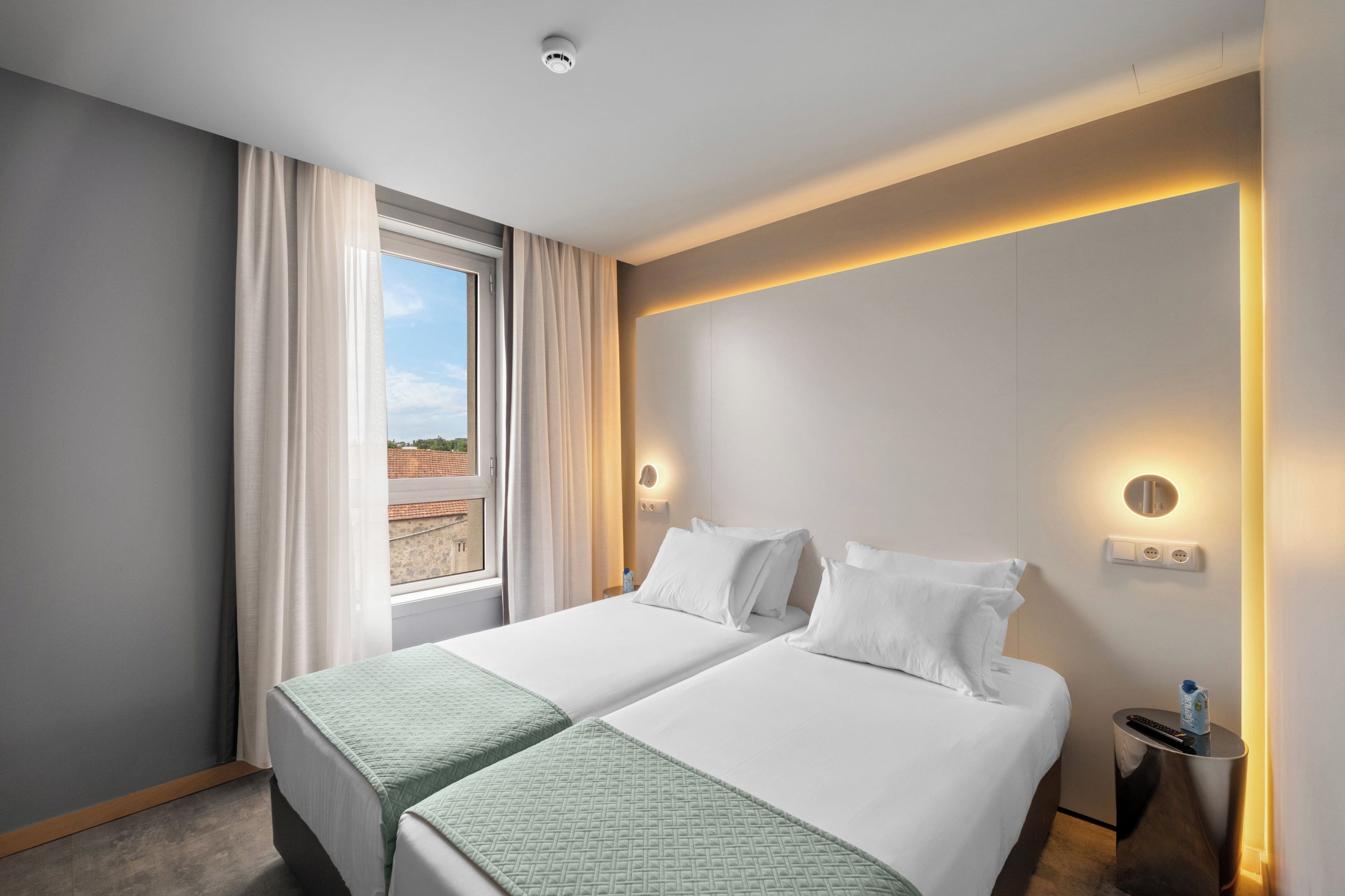 Een hotelkamer met twee bedden, een raam met gordijnen, een nachtkastje met een lamp en een wandlamp.