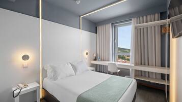 Quarto de hotel moderno com cama, mesa de cabeceira com telefone, escrivaninha com cadeira, janela com cortinas e guarda-roupa embutido.